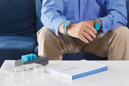 A man sits on a sofa, wearing monitoring devices on his wrists.